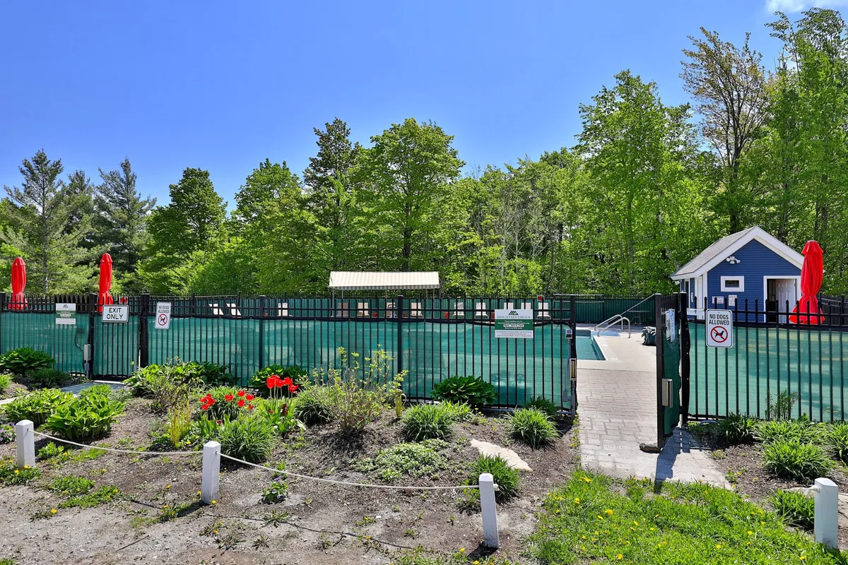 An inviting outdoor swimming pool surrounded by a landscaped garden and an entry gate, with lounge spaces for sunbathing and gathering. The clear water and lush greenery evoke the vibrant energy of summer activities.