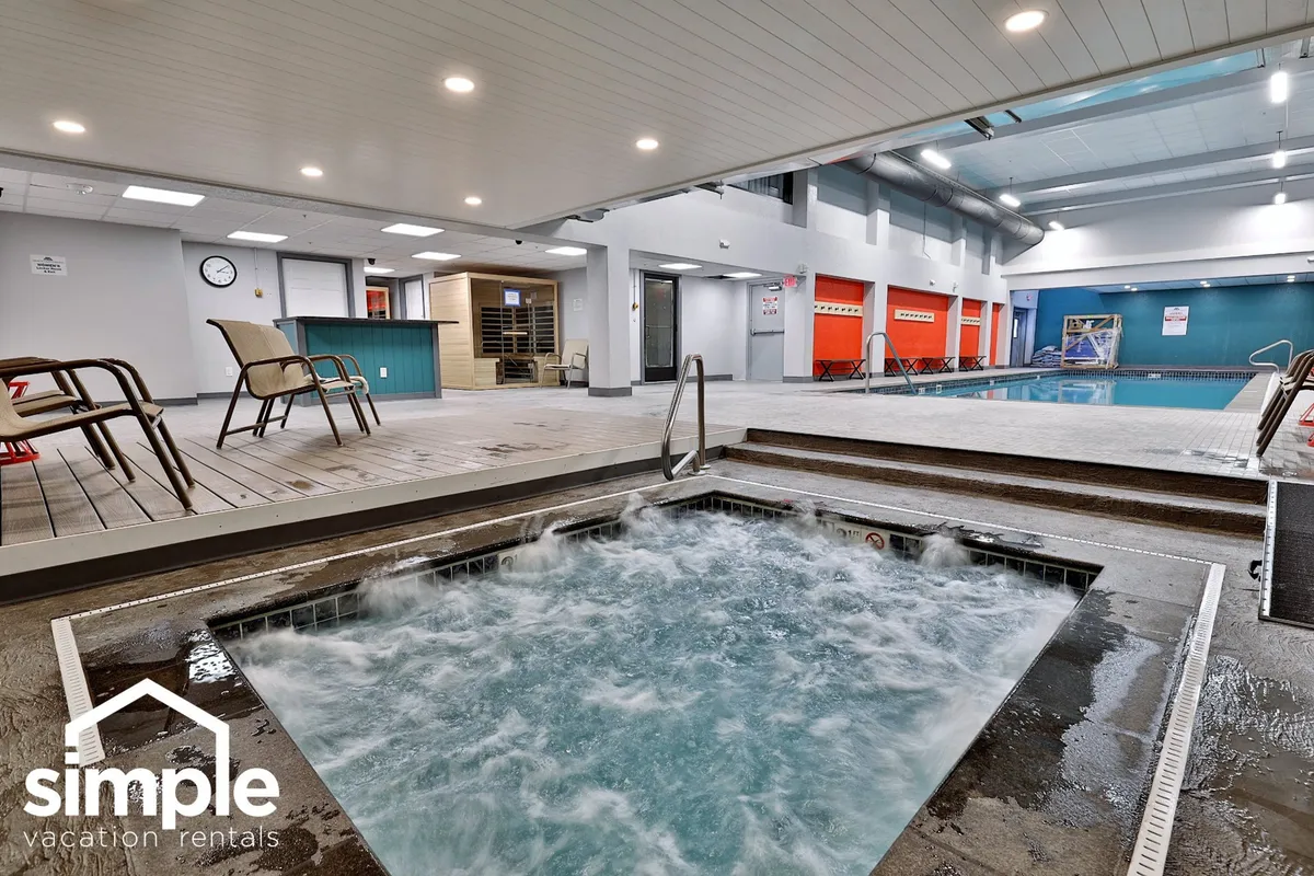 A tranquil indoor pool area featuring a large swimming pool, adjacent hot tub, and comfortable lounge chairs. The space is bathed in soft lighting, inviting guests to relax, swim, or soak after a day on the slopes. A clock and windows add a sense of openness and time, perfect for unwinding at any hour.