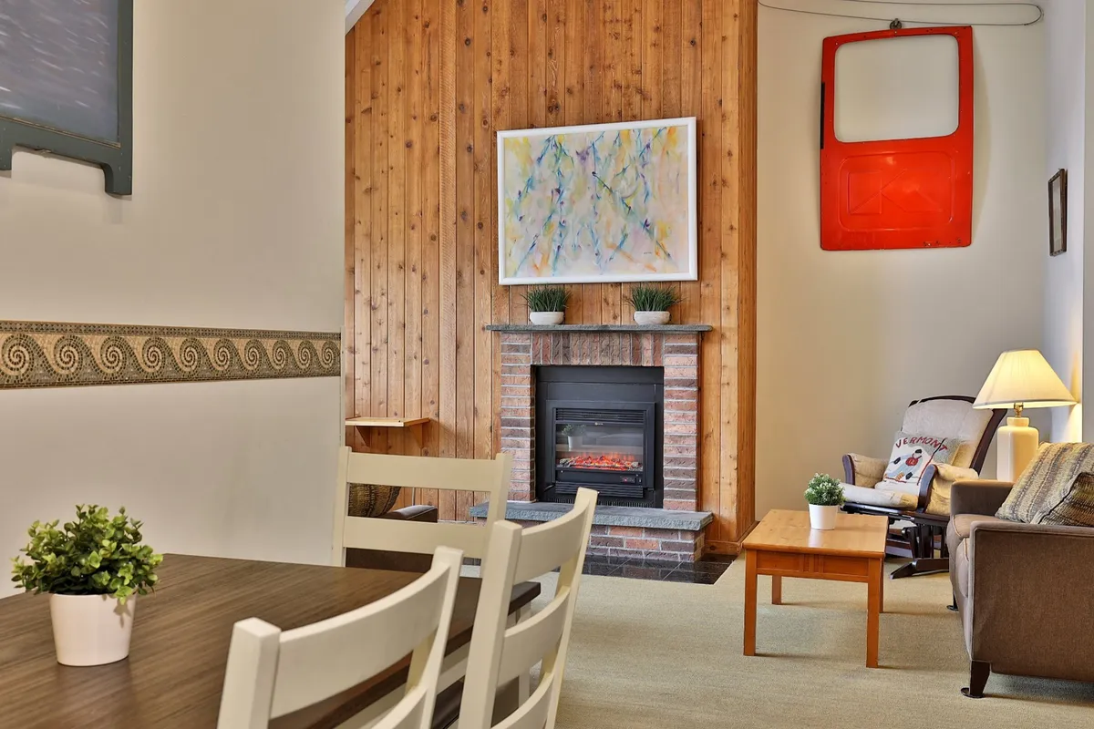 A cozy dining space featuring a fireplace, wood paneling, artwork, and a well-set dining table. The ambiance is inviting, with the fireplace suggesting warmth and relaxation, perfect for extended meals and evenings indoors.