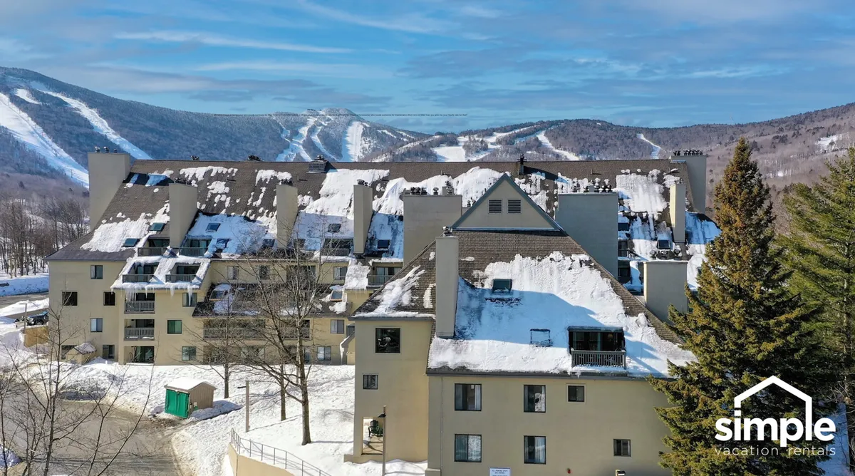 A scenic exterior view featuring snow-covered rooftops set against a stunning mountain backdrop, framed by pine trees. The scene captures the quintessential après-ski atmosphere, evoking both the thrill of the slopes and the warmth of returning home.