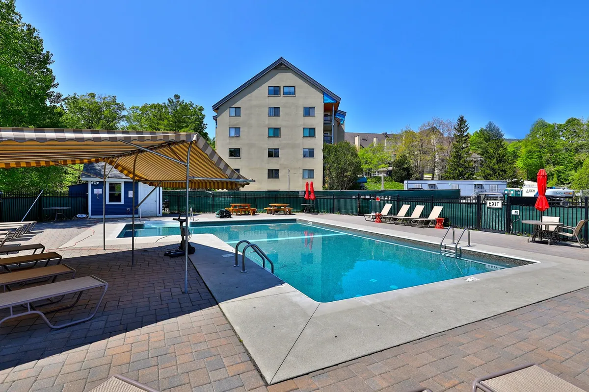 A vibrant outdoor swimming pool surrounded by lounge chairs, umbrellas, and lush greenery. The space is perfect for summer relaxation, socializing, or evening gatherings by the water.