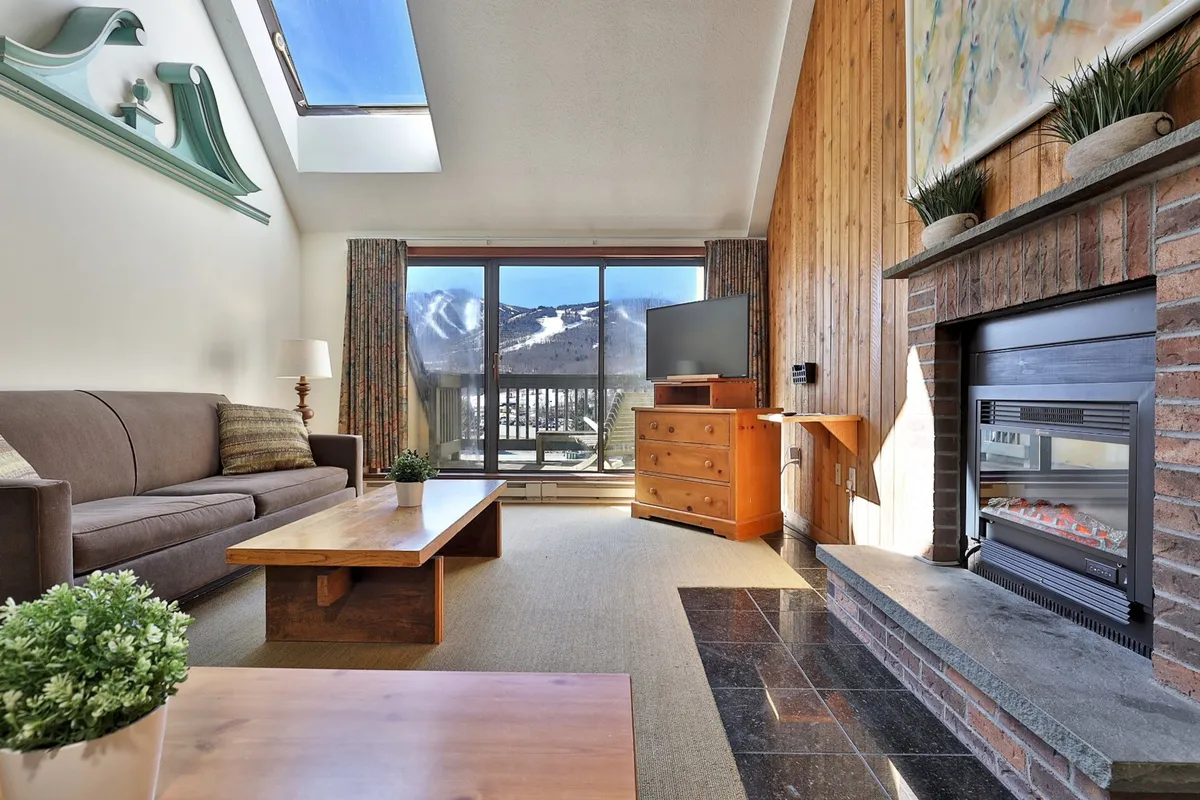 A bright, comfortable living room with a plush sofa, modern fireplace, large windows offering mountain views, and a coffee table at the center. The ambiance is tranquil, inviting relaxation, socializing, or quiet evenings by the fire. The window creates a strong visual connection to the outdoors, reflecting the mountain setting.