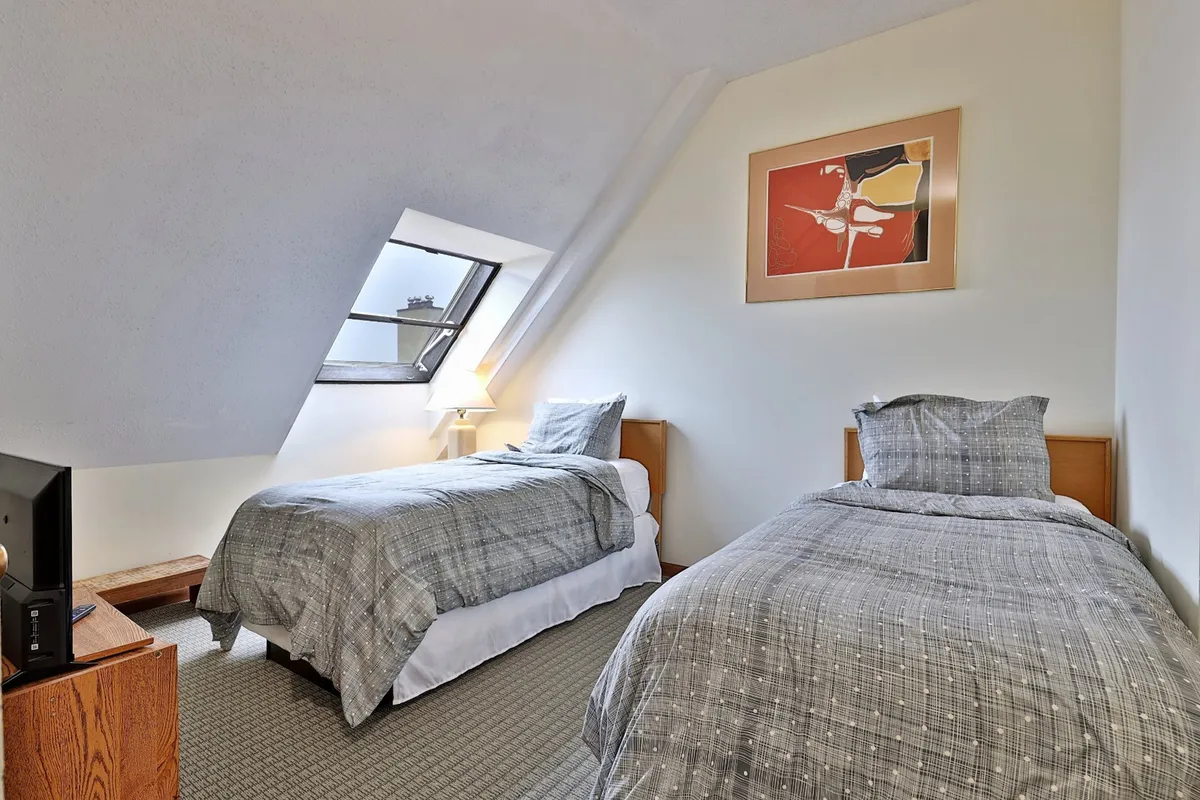 A tranquil bedroom with two single beds, simple artwork on the wall, a window bringing in natural light, and a television. The layout is open and inviting, ideal for children or friends.