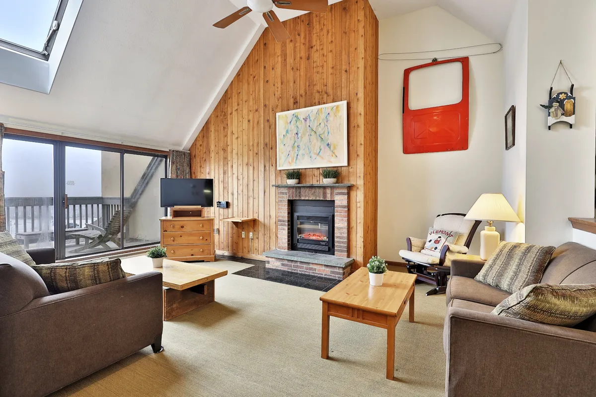 A warm, inviting living room featuring a wooden accent wall, a roaring fireplace, large windows framing scenic views, and coffee tables positioned for intimate conversation. The cozy setup encourages relaxation with plush seating, ambient lighting, and tasteful artwork.