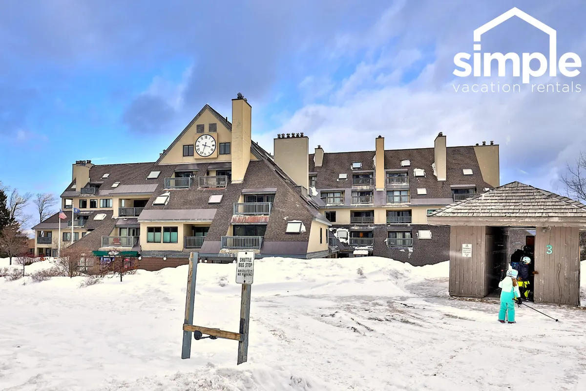 The exterior of a ski lodge with a prominent clock, snowy landscape, and guests—some carrying skis—gathering near the entrance. The scene conveys both the lively energy around the base area and the welcoming, cozy vibe of returning after a day on the mountain.